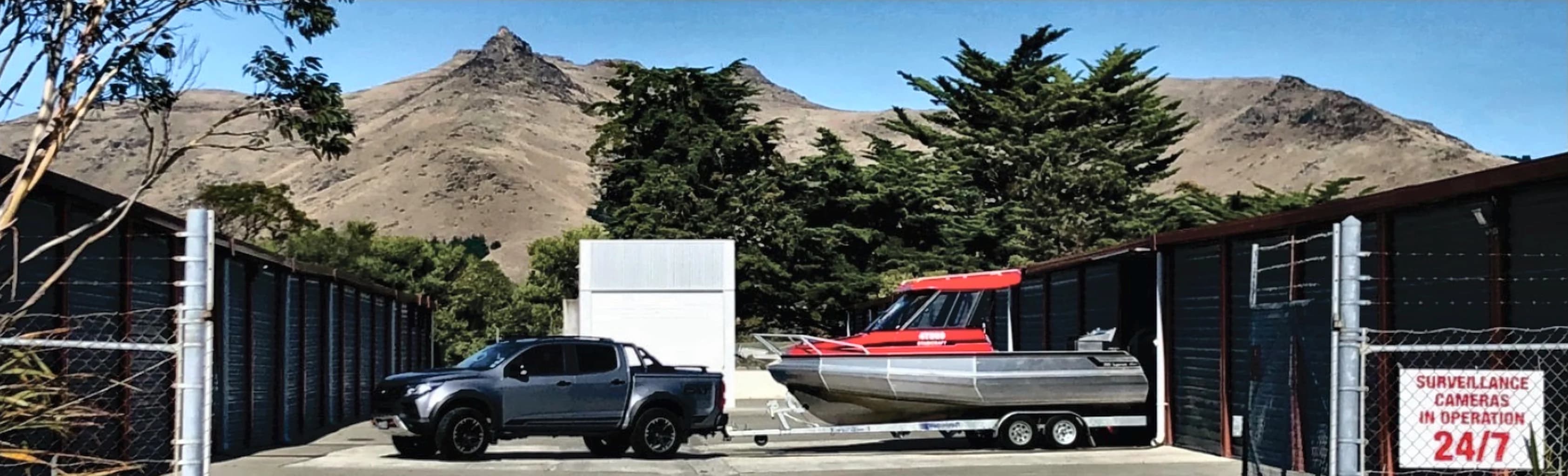 A ute towing a boat inside the Ferrymead Storage yard, with the units on both sides and the Port Hills behind